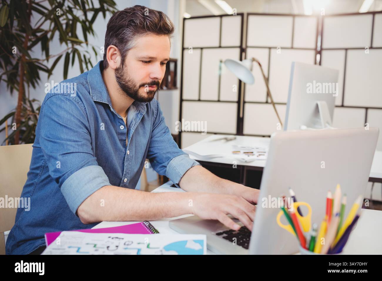 Anfang der 30er Jahre tippte ein Mann auf einem Laptop in einem modernen Büro, mit Skizzen und Pflanzen, Kopierraum Stockfoto