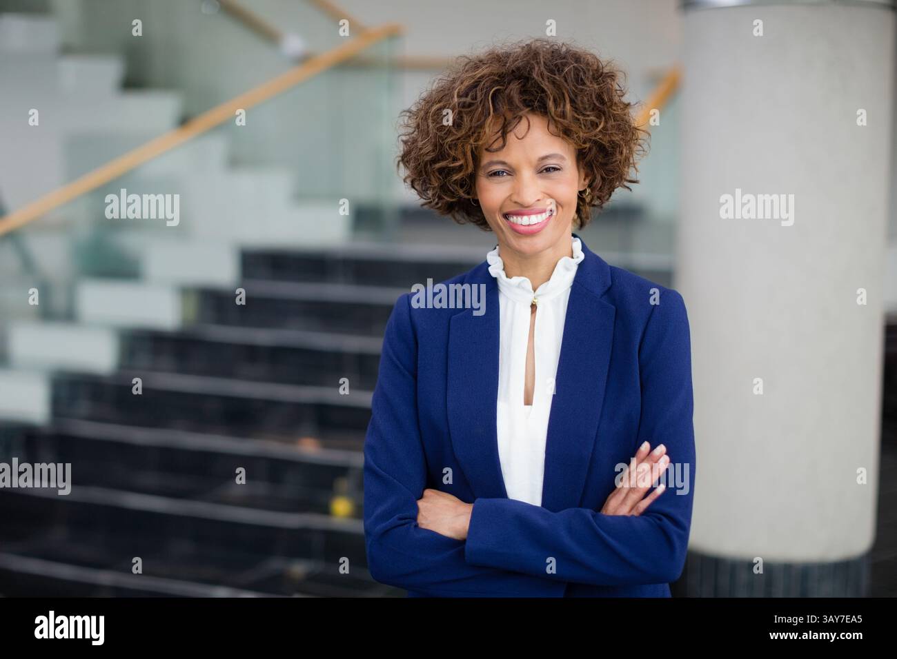 Afroamerikanische Frau, die mit gefalteten Armen in der Firmenlobby an einer Glastreppe steht, Kopierraum Stockfoto