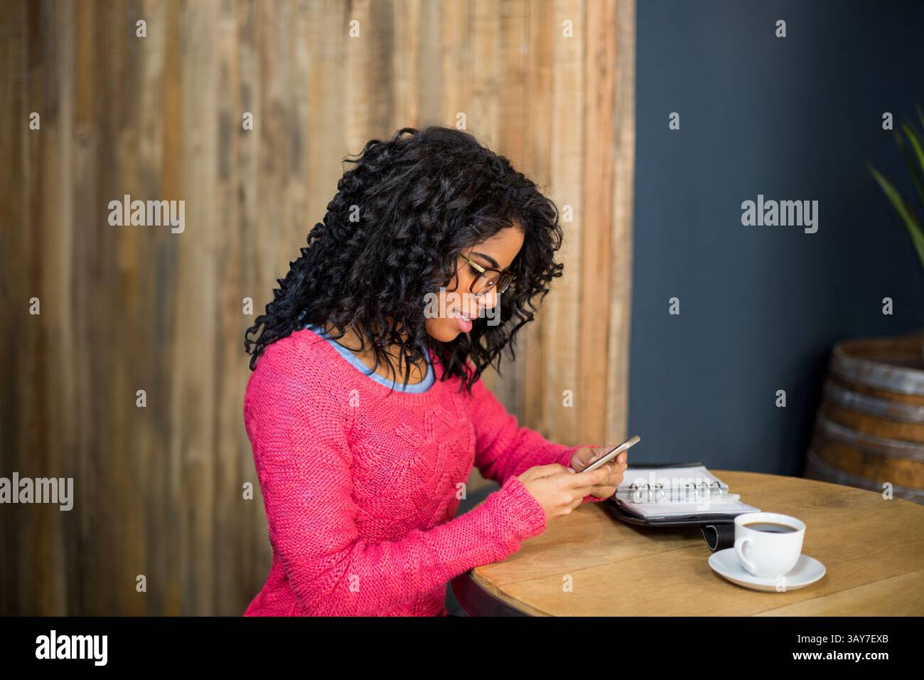 Frau in rosa Pullover sitzt am Tisch im Café und schaut auf Smartphone mit Planer und Kaffee Stockfoto
