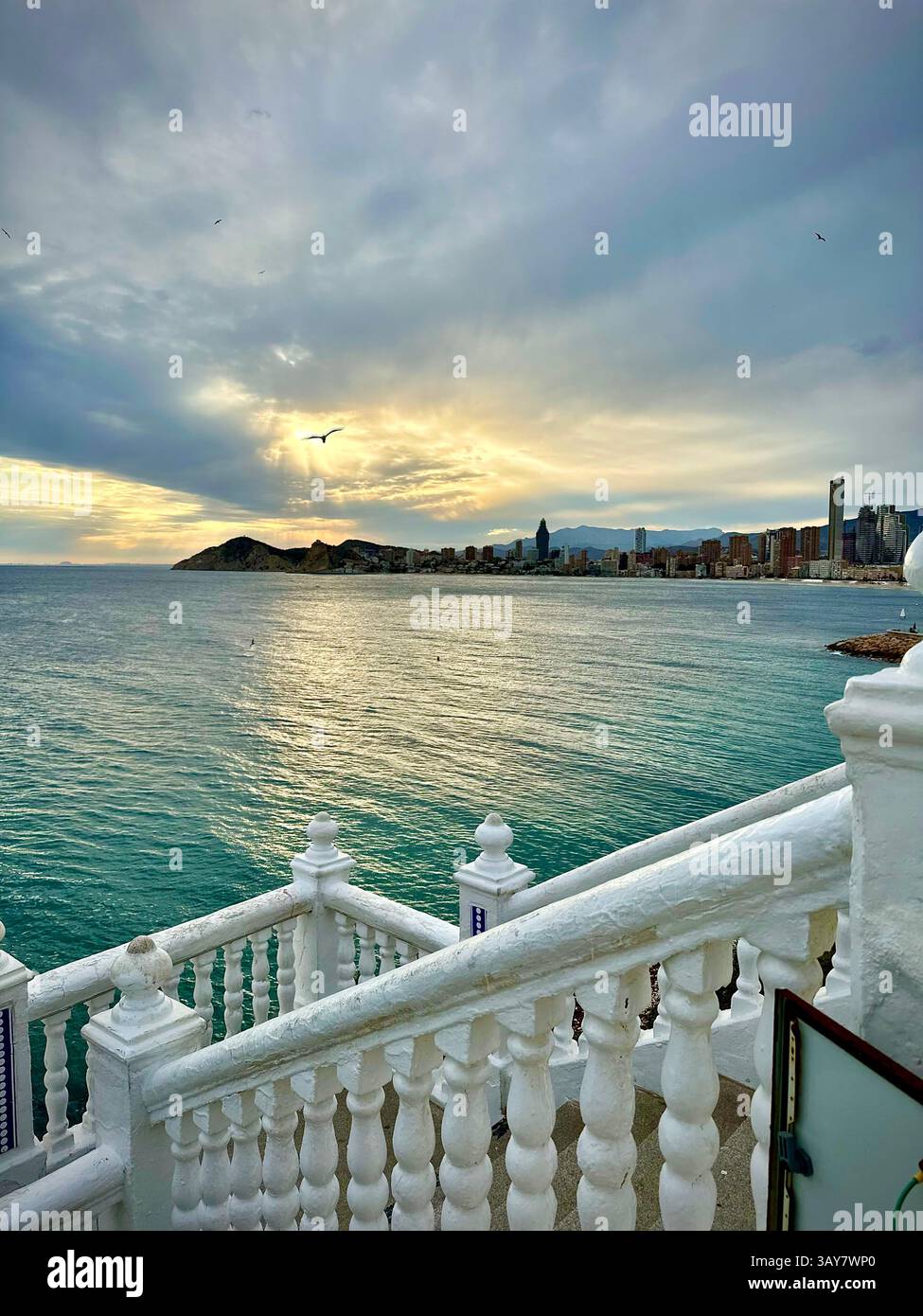 Eine weiße Balustrade umrahmt einen atemberaubenden Sonnenuntergang über einer Skyline der Küste, mit sanften Wellen und warmem Licht, die einen friedlichen Ton bilden. Die Szene fängt das e ein - Smartphone-aufgenommenes Stockfoto