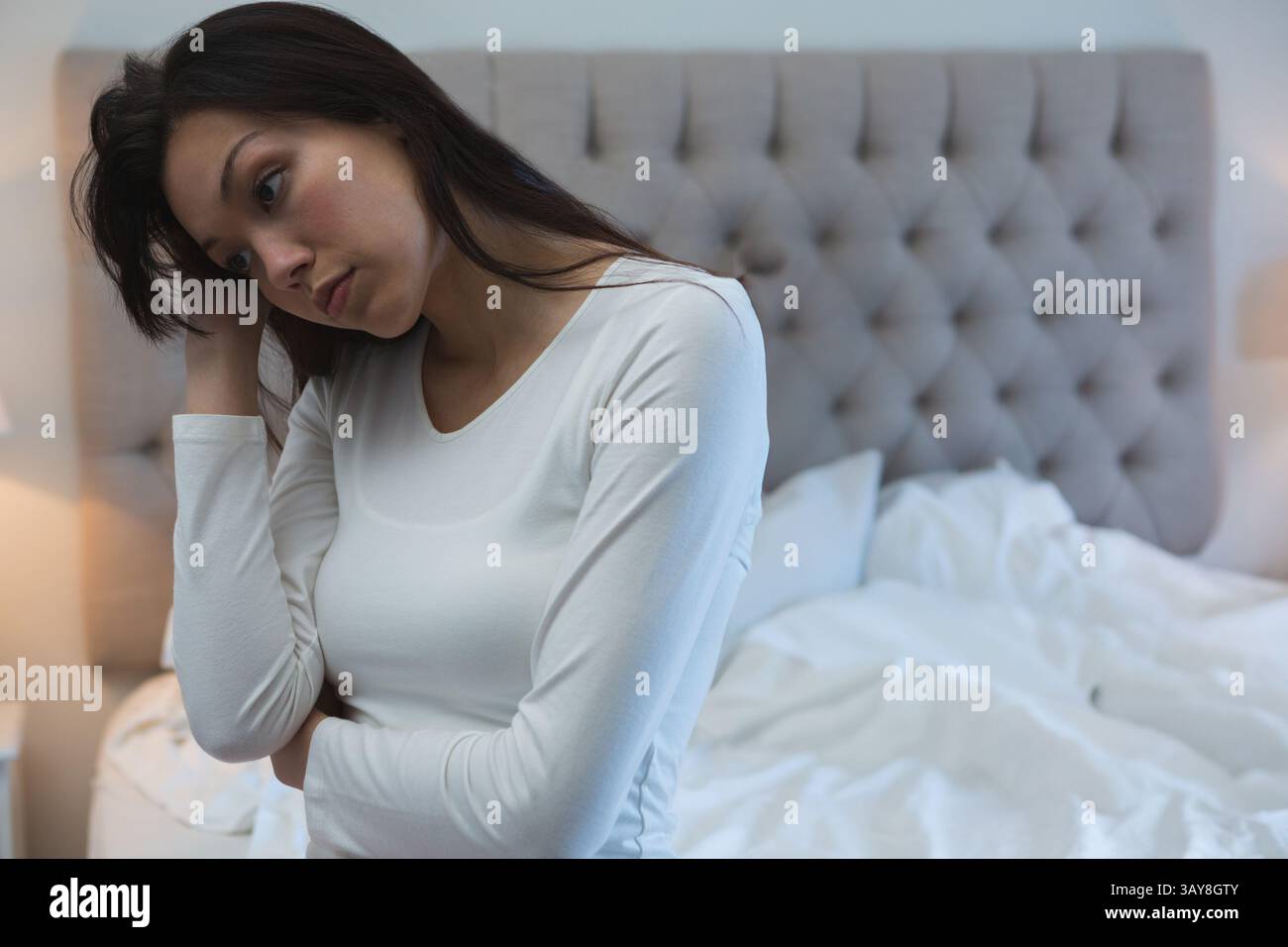 Junge asiatische Frau, die sich im Schlafzimmer mit dem Kopf auf die Hand lehnt, nachdenklich mit grauem Kopfteil, Kopierraum Stockfoto