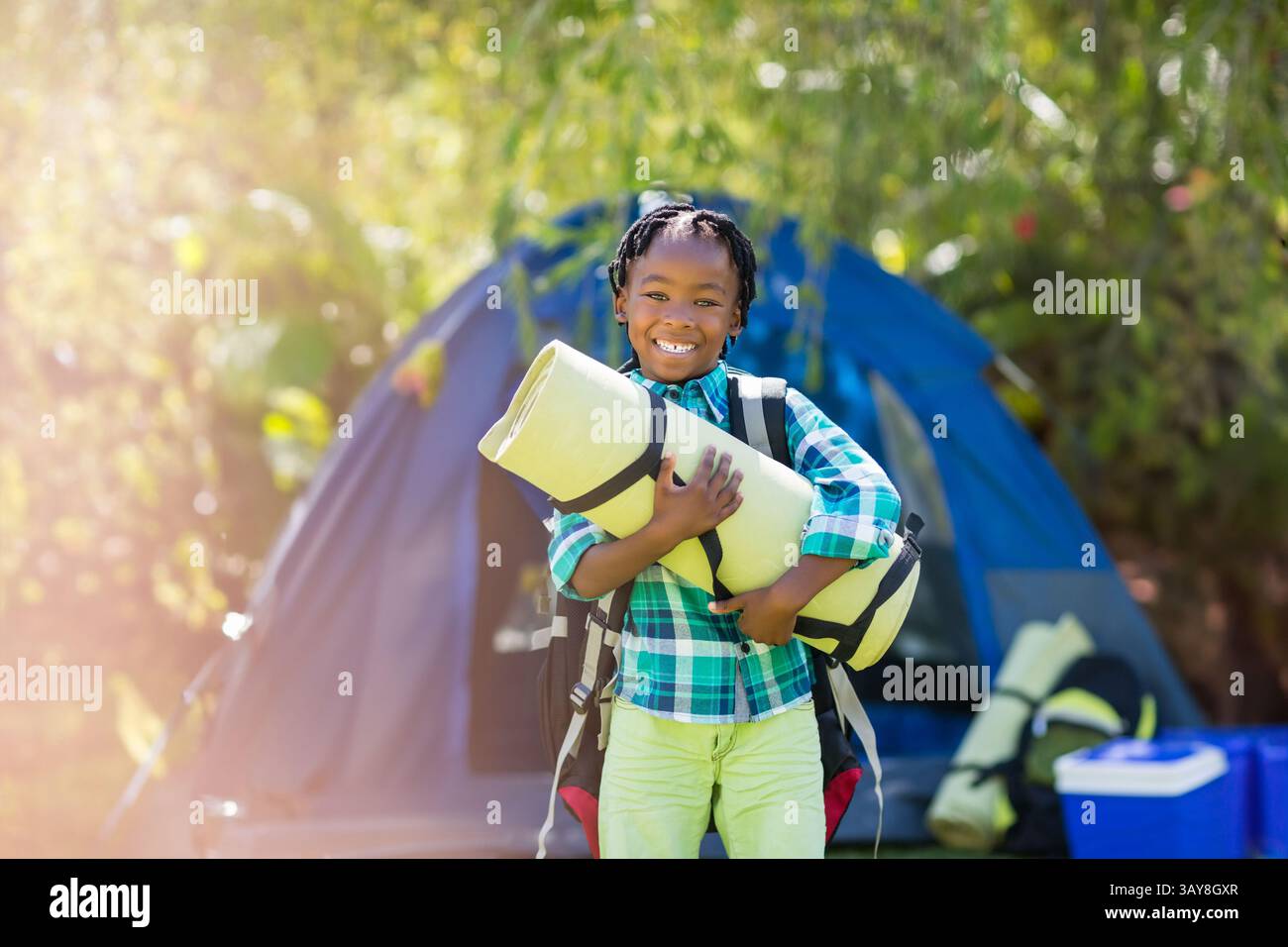 Afroamerikanerin, die auf dem Waldcamping steht, eine Matte hält, einen Rucksack mit Kühlbox trägt Stockfoto