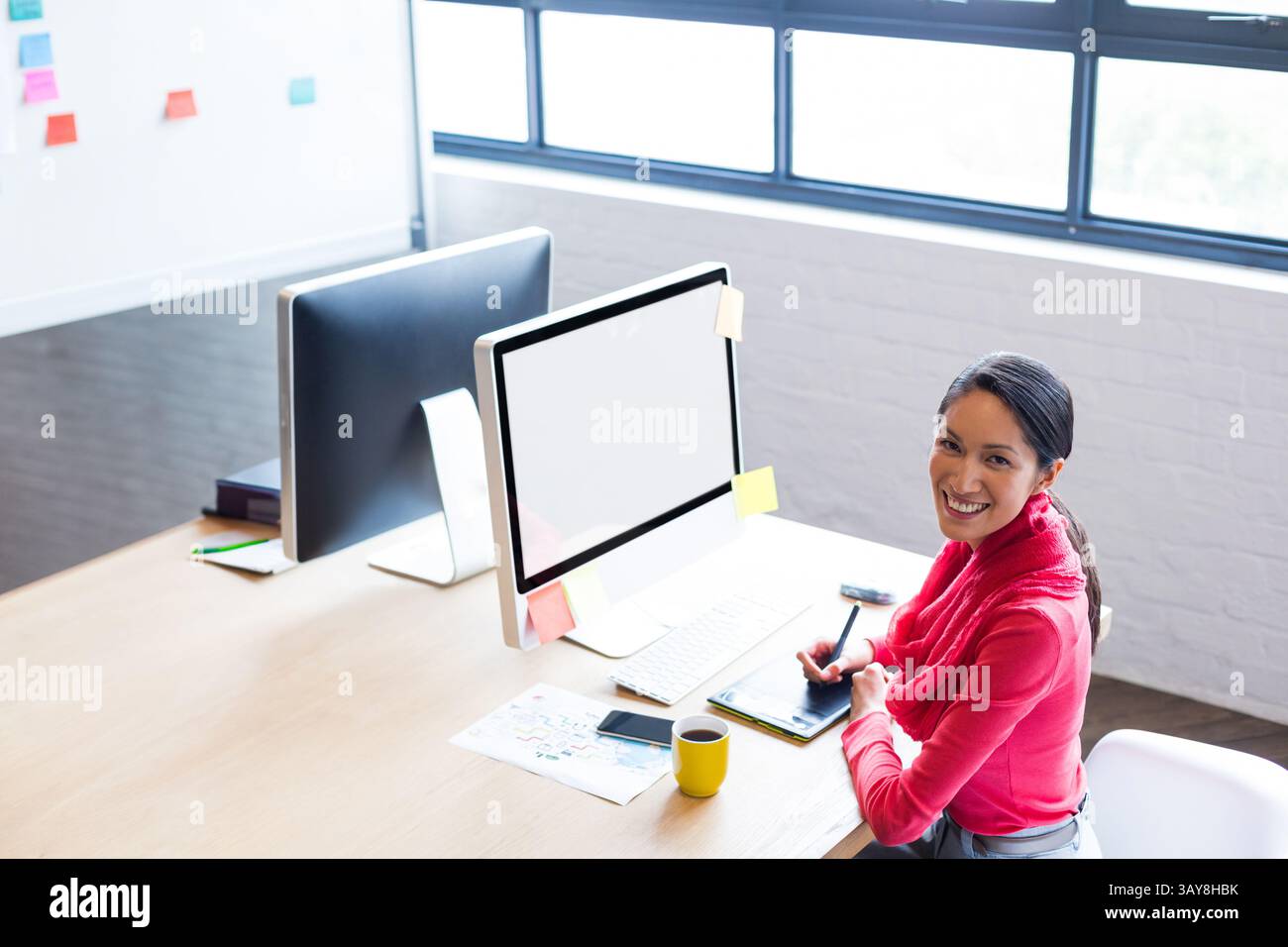 Junge Chinesin, die im Büro Kunst auf Zeichentablett erstellt, mit zwei Monitoren und Haftnotizen Stockfoto