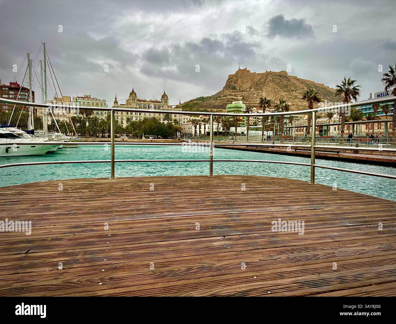 Malerische Perspektive von einer hölzernen Promenade mit Blick auf einen türkisfarbenen Hafen voller Yachten, eingerahmt von Palmen und historischer Architektur. Ein roc - Smartphone-aufgenommenes Stockfoto