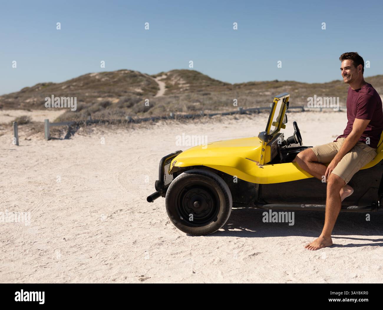 Mann, der barfuß auf gelbem Dünenwagen in Küstendünen sitzt, mit Holzpfosten, Kopierraum Stockfoto