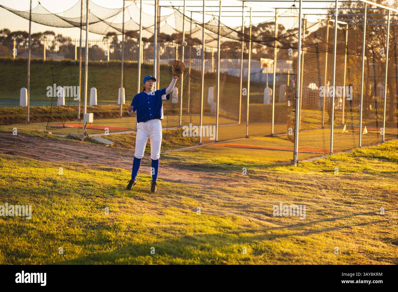 zweirassige junge Frau, die auf dem Grasfeld neben Schlagkäfigen Ball fängt, mit Lederhandschuh Stockfoto