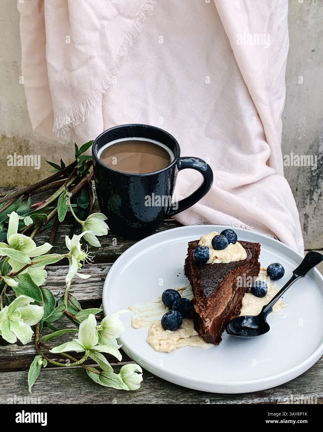 Ein Stück reichhaltiger Schokoladenkuchen mit Heidelbeeren und Sahne, serviert auf einer Keramikplatte neben einer schwarzen Tasse Kaffee. Stockfoto