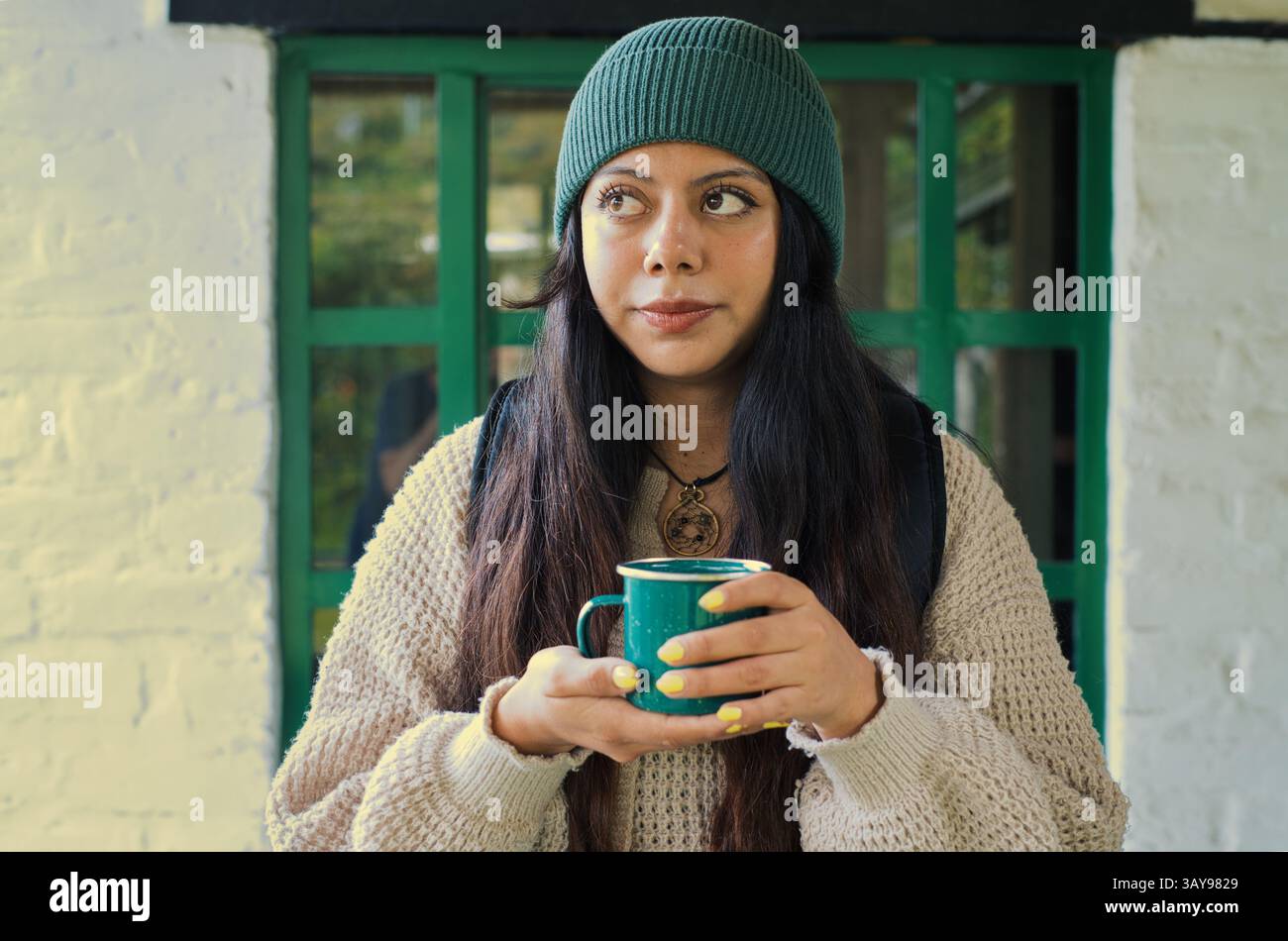Eine junge Frau mit einer stilvollen Mütze hält eine warme Tasse Kaffee und hält einen ruhigen Moment in einer gemütlichen Umgebung fest Stockfoto