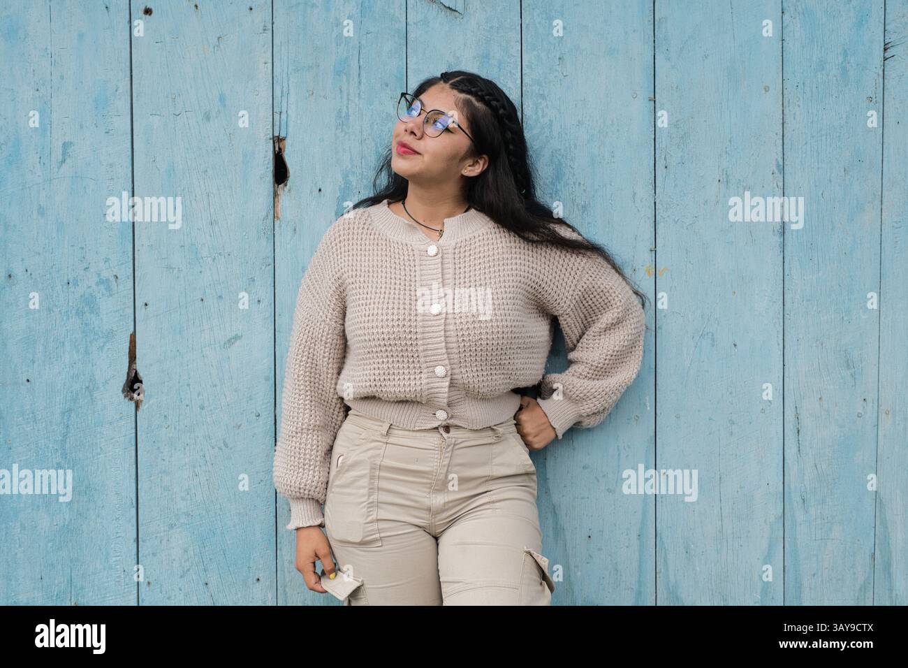 Eine stilvolle und modische Frau, die eine Brille trägt, posiert selbstbewusst neben einer leuchtend blauen Holzwand Stockfoto