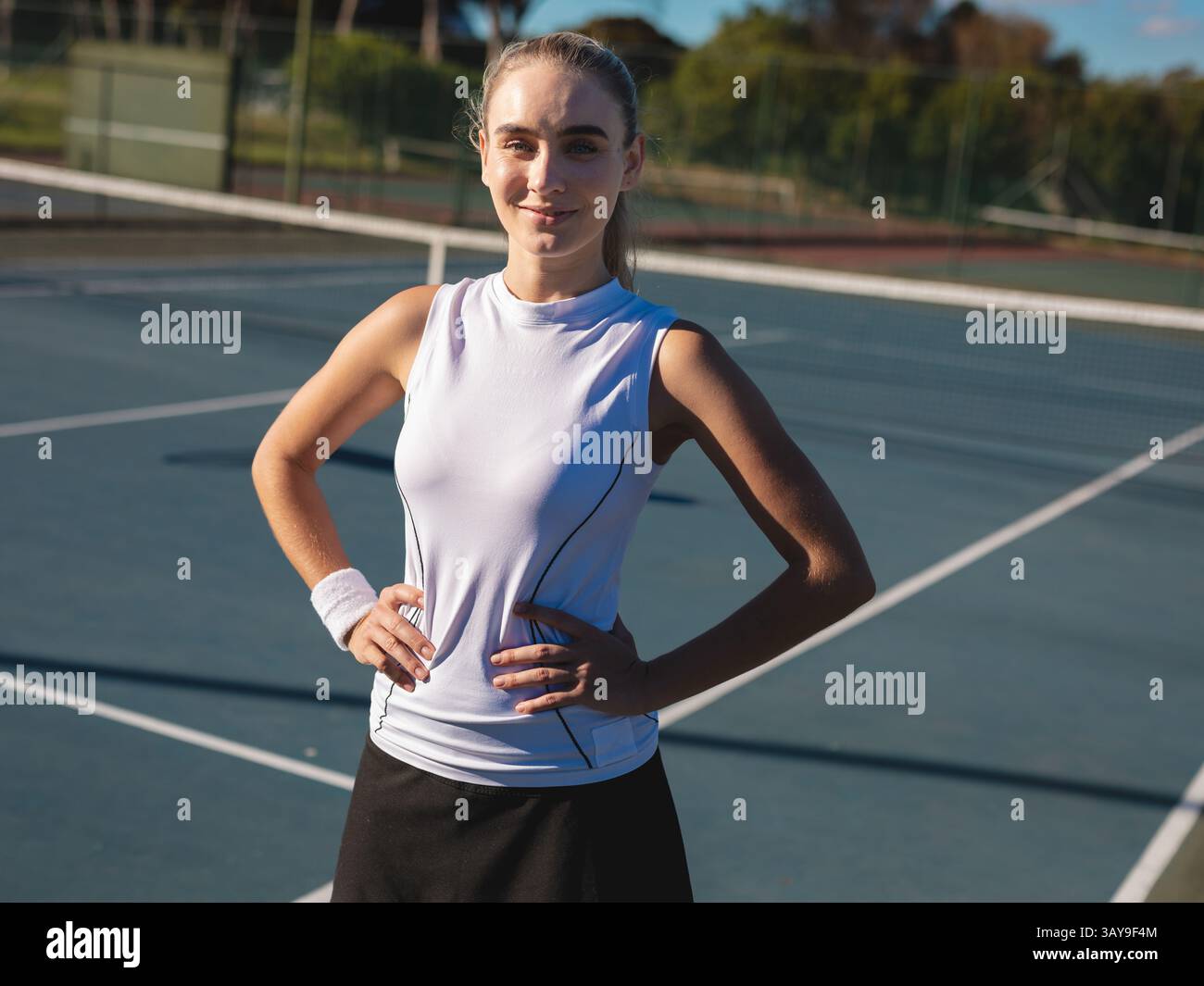 Junger erwachsener Tennisspieler, der mit Händen auf Hüften auf dem Hartplatz steht, Netz und Armband zeigt Stockfoto