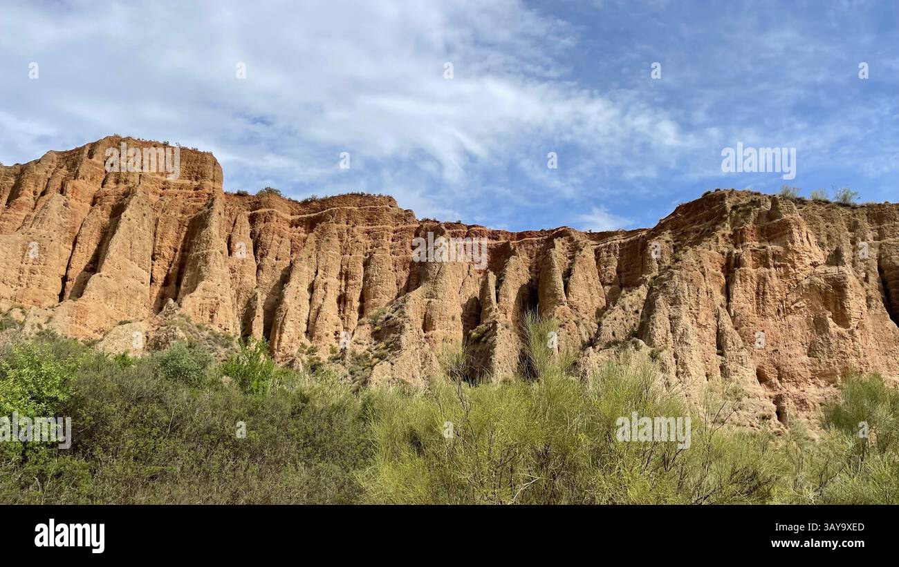 Erodierte Landschaft in Alpujarra, Granada, Andalucía, Spanien mit Pflanzen im Vordergrund und einem wolkenblauen Himmel darüber. Stockfoto