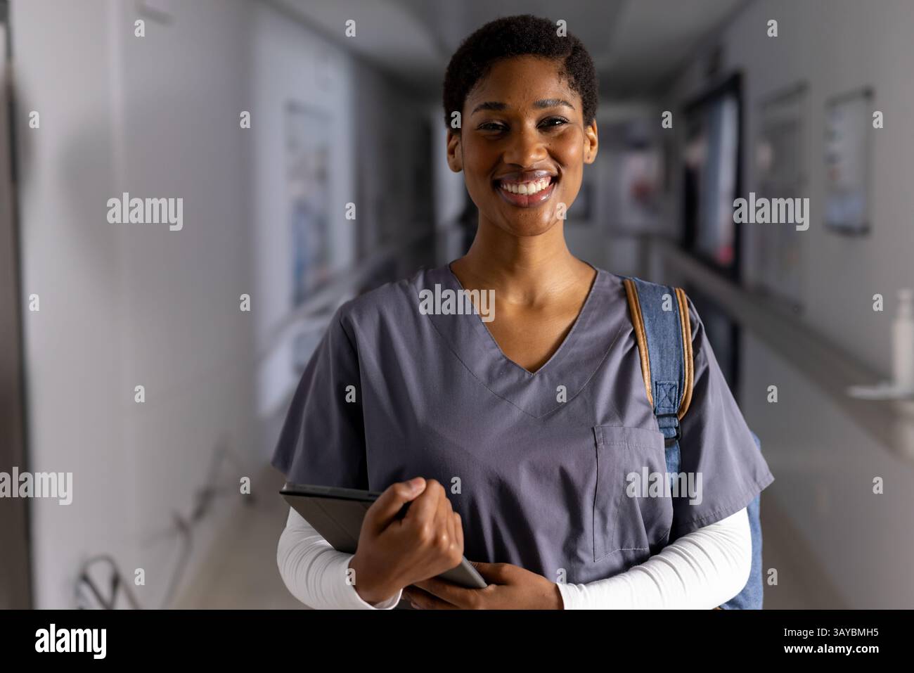 Afroamerikanerin, die im Krankenhausflur steht, Tablette hält und einen Rucksack trägt, lächelt Stockfoto