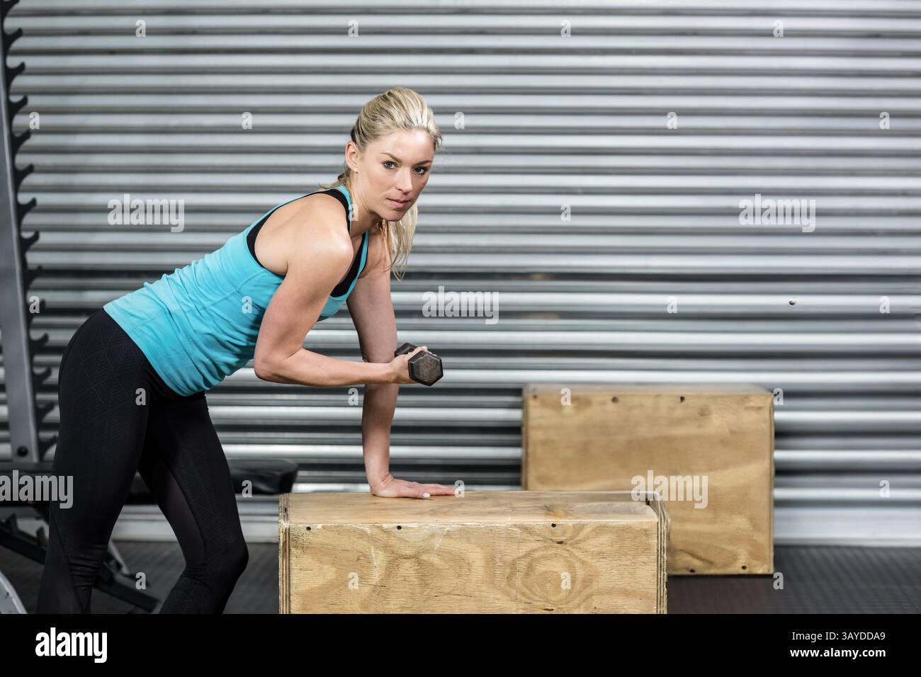 Junge Erwachsene Frau, die sich auf eine hölzerne Plyometrische Box lehnt und die Hexenhantel im Fitnessstudio hebt, zeigt Stärke Stockfoto