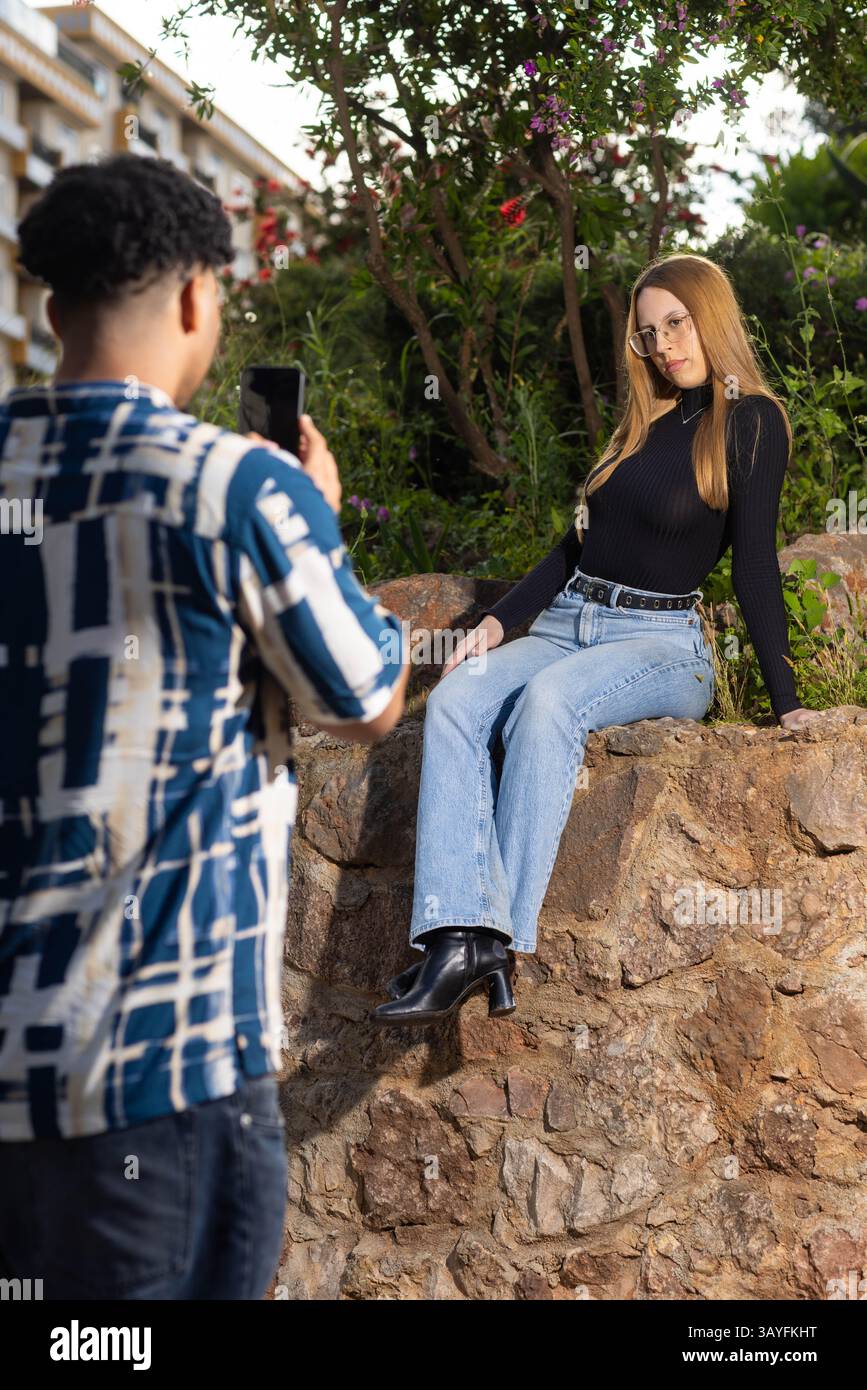 Junge Frau posiert auf einer Steinmauer, während sie draußen in einem zwanglosen Urban Garden Fotoshooting die Kamera anlächelt Stockfoto