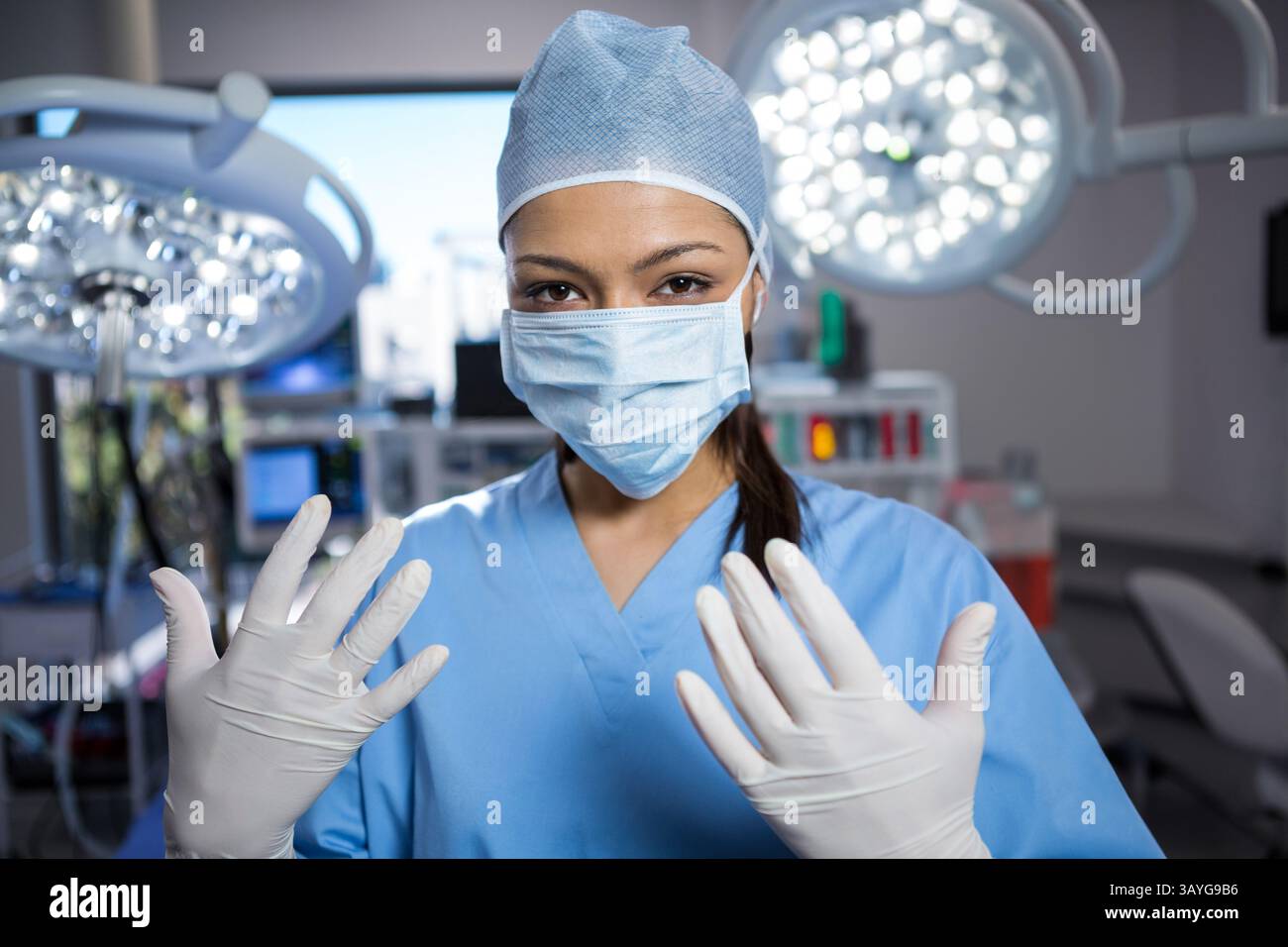Weibliche Chirurgin, die Handschuhe und Scheuerkappe im Operationssaal mit Deckenleuchten und Monitoren anbringt Stockfoto