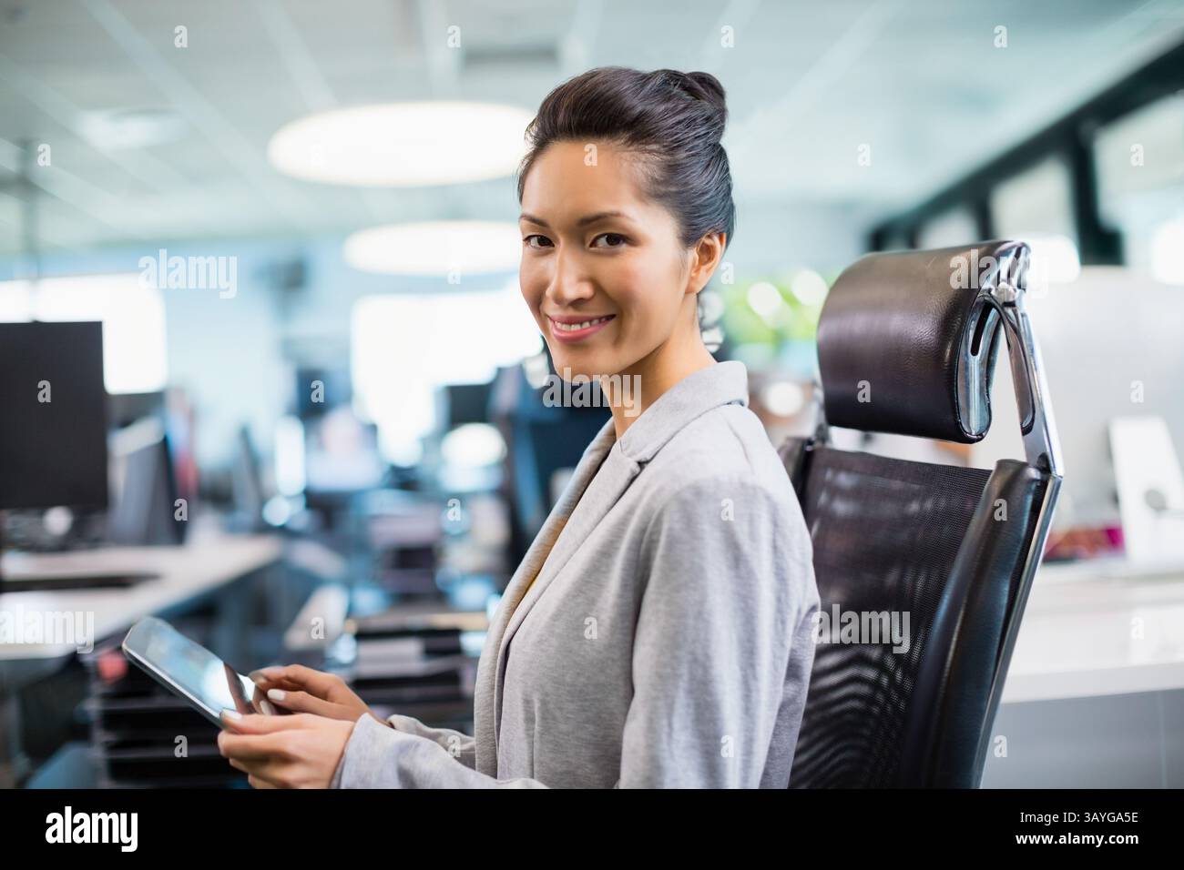 Asiatische Frau, die ein Tablet hält und in einem ergonomischen Stuhl im Großraumbüro sitzt, mit Kopierraum Stockfoto