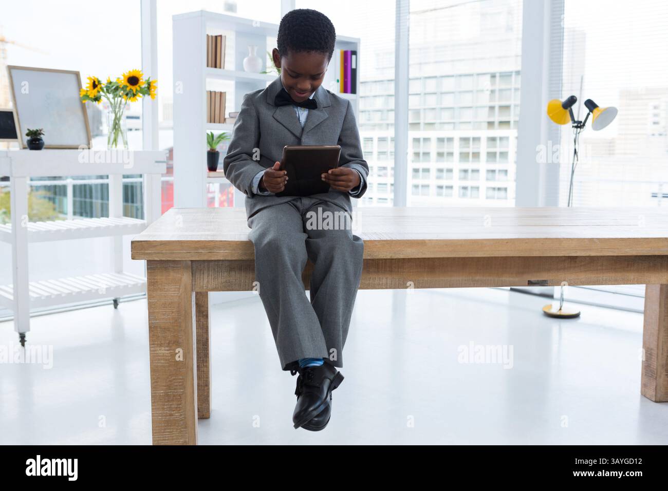 Afroamerikanischer Junge, der auf einem Tisch in einem modernen Studio sitzt und ein Tablett in der Nähe von Sonnenblumen hält Stockfoto