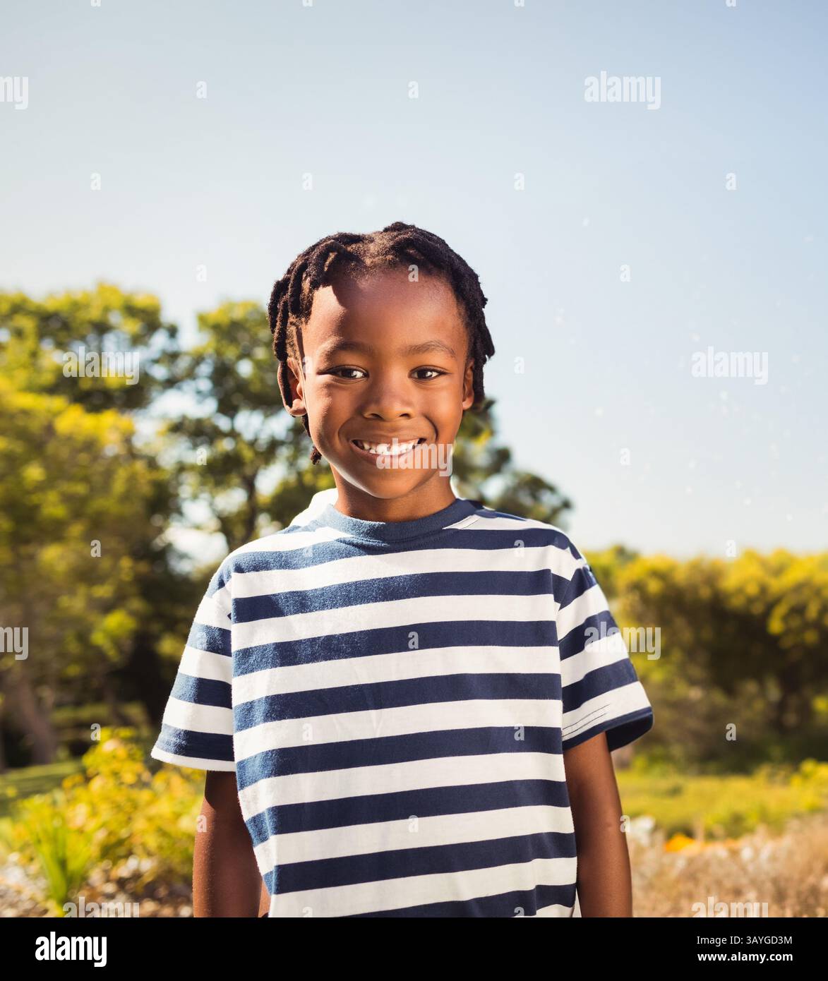 Ein afroamerikanischer Junge, der im sonnigen Park steht und lächelt, trägt ein marineblaues und weiß gestreiftes T-Shirt Stockfoto