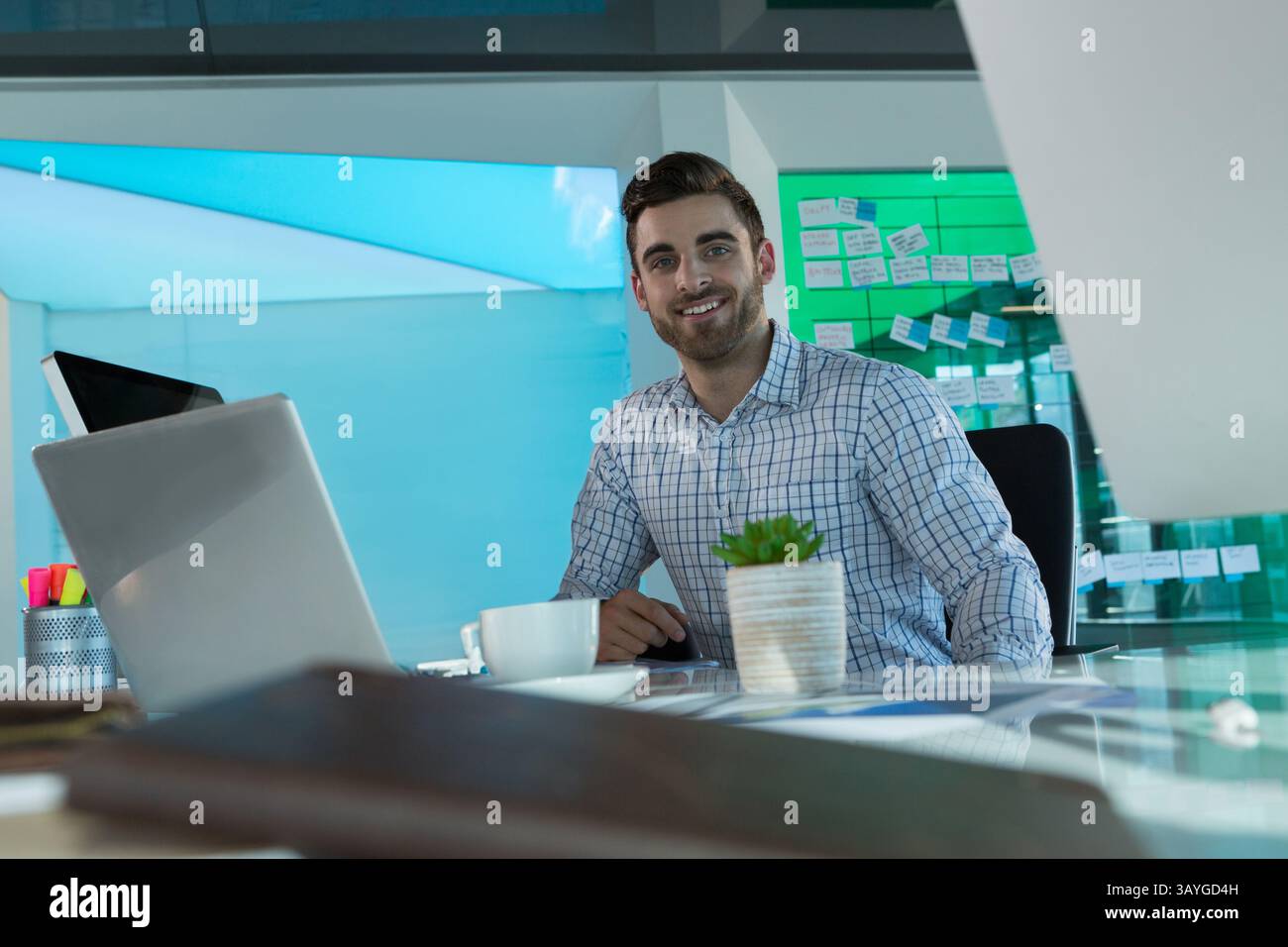 Mann hält Stift und lächelt am Glasschreibtisch im Arbeitsbereich mit Laptop, Kaffeetasse, Stifthalter Stockfoto