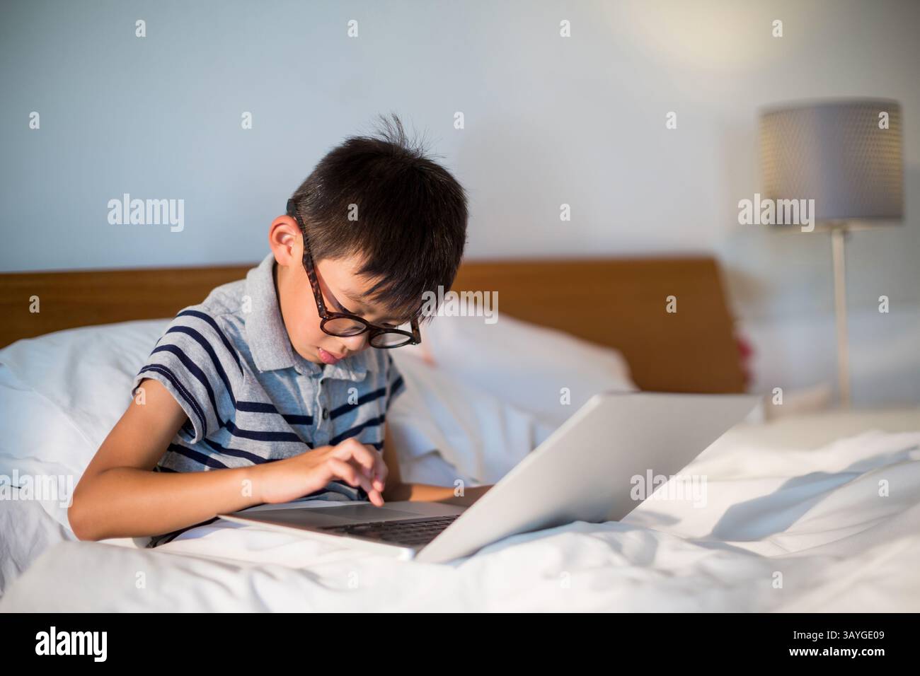 Asiatischer Junge, der im Schlafzimmer mit Brille und Lampe auf dem Bett sitzt, mit silbernem Laptop Stockfoto