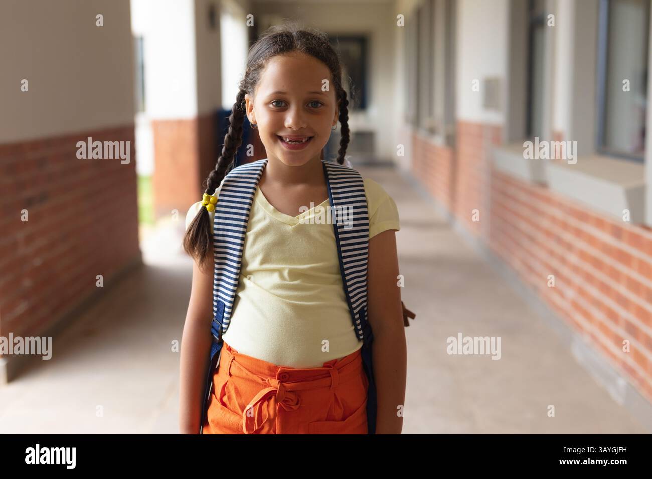 Mädchen, das im Schulflur steht und lächelt, trägt einen gestreiften Rucksack und gelbe Haarspangen Stockfoto