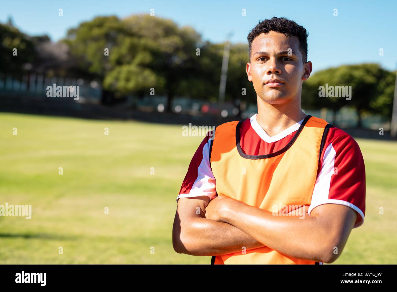 Männlicher Athlet, der auf dem grasbewachsenen Sportplatz steht, trägt ein orangefarbenes Lätzchen mit rot-weißem Jersey, Kopierraum Stockfoto