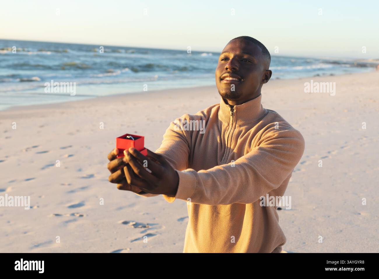 Afroamerikaner kniet am Sandstrand, präsentiert rote Box mit Diamantring, Kopierraum Stockfoto