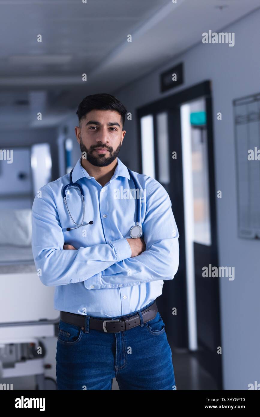 Indischer Arzt, der mit gekreuzten Armen in der Krankenhaushalle steht, mit Stethoskop, Trage Stockfoto