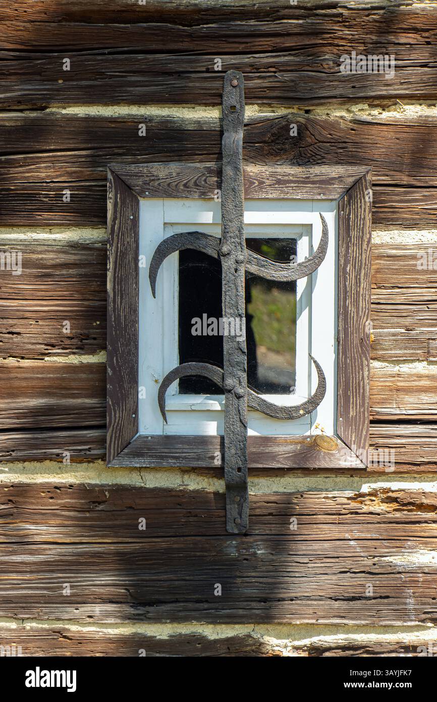 Kleines Fenster an der Wand eines alten Holzhauses mit einem schmiedeeisernen Grill. Stockfoto