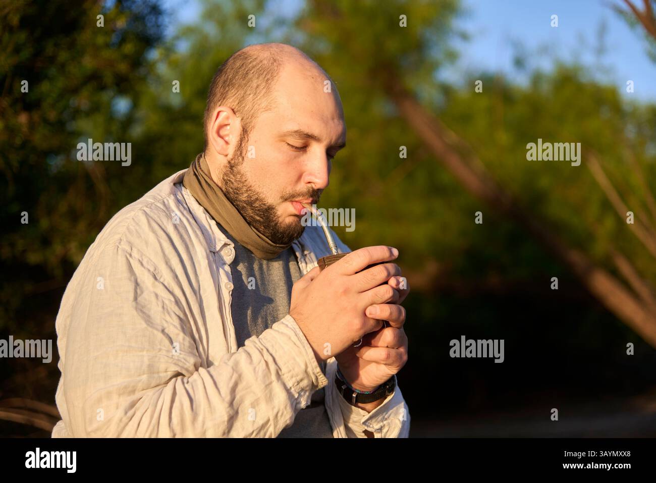 Hispanischer Reisender, der bei Sonnenaufgang am Ufer eines Baches sitzt und die ersten Sonnenstrahlen empfängt, während er Mate trinkt, ein typisches argentinisches Getränk. El Stockfoto