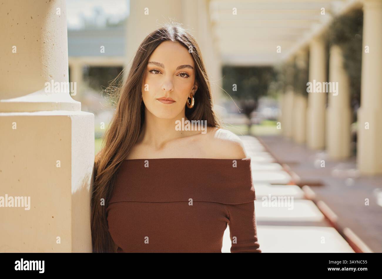 Eine junge Frau mit langen braunen Haaren, die ein mokkafarbenes, schulterfreies Rippenoberteil trägt, posiert anmutig neben eleganten Säulen an einem sonnigen Tag Stockfoto