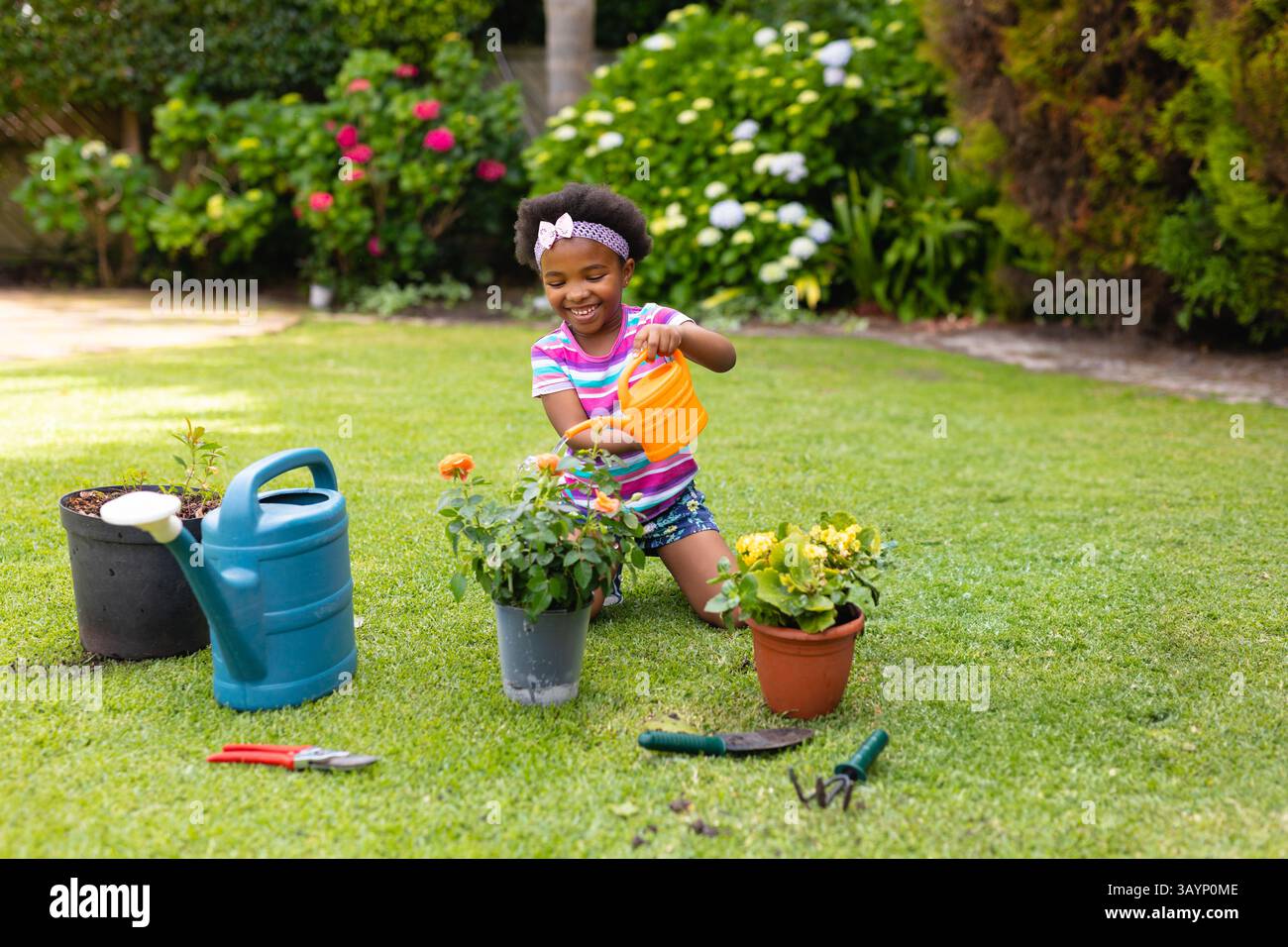 Afroamerikanisches Mädchen kniet und bewässert Pflanzen im Garten des Hinterhofs, mit Gießkannen und Werkzeugen Stockfoto