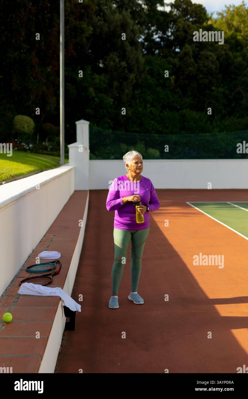 Seniorin steht auf einem privaten Tennisplatz im Freien und hält eine Wasserflasche mit Schlägern und Handtuch Stockfoto