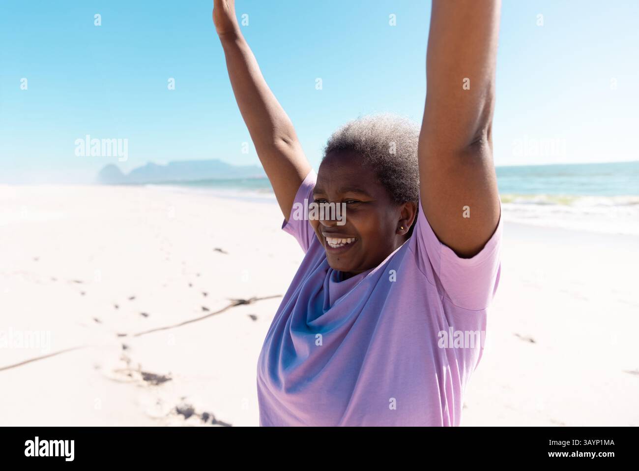 Afroamerikanerin feiert auf dem Meer am sonnendurchfluteten Sandstrand mit Lavendel-T-Shirt Stockfoto