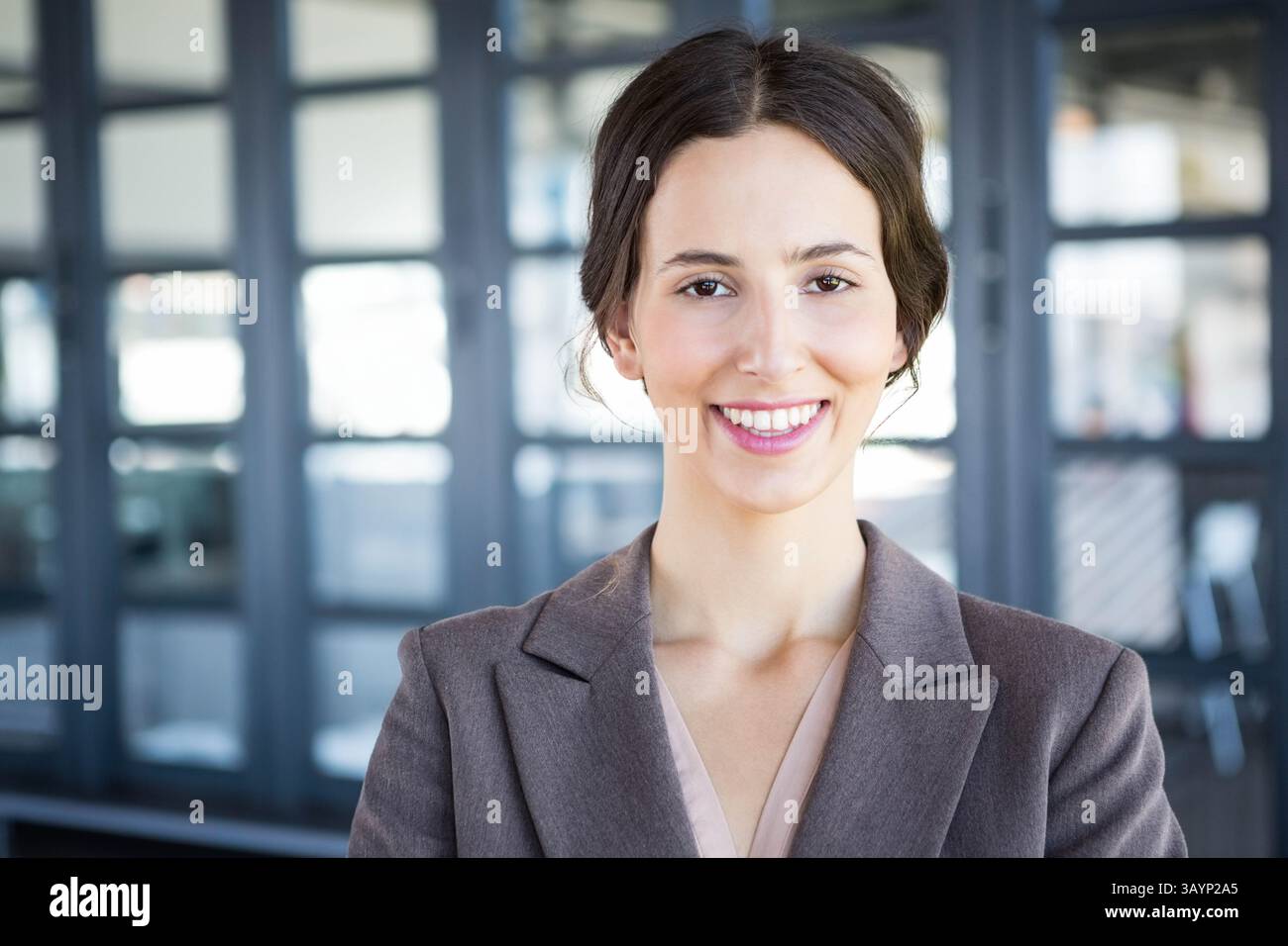 Frau lächelt, während sie im Büro steht, mit Glaswänden, grauem Blazer und leichter Bluse Stockfoto