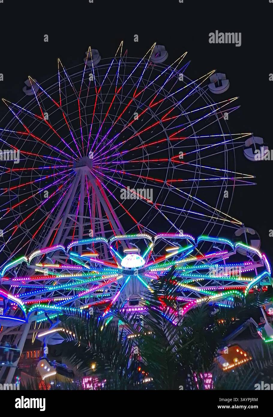 Pulsierende Nachtszene in einem Vergnügungspark mit einem großen Riesenrad, das von bunten Lichtern und Karussells in hellen Neontönen beleuchtet wird. Festliche Atmosp Stockfoto