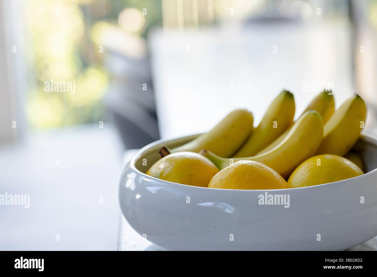Die weiße Keramikschale hält Bananen und Orangen auf der Tischplatte in der modernen Küche nahe dem sonnendurchfluteten Fenster Stockfoto