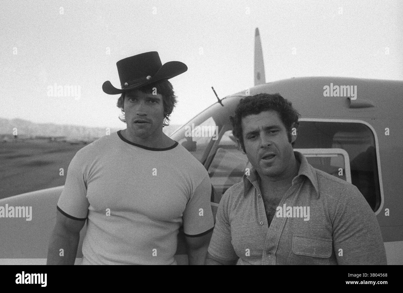 Juli 1979; Los Angeles, CA, USA; der professionelle Bodybuilder ARNOLD SCHWARZENEGGER trägt einen Cowboyhut und posiert für ein Foto mit seinem Freund BILL DRAKE und seinem Flugzeug. Arnold, geboren in einem kleinen Dorf in Österreich, nahm schon in jungen Jahren das Krafttraining auf und stieg bald als Bodybuilding-Champion an die Spitze. Sein Einbruch in der Schauspielerei kam 1982 in dem Film Conan the Barbarian. Seit Conan hat Arnold eine lange Karriere als Schauspieler. Arnold war seit Anfang der 80er Jahre in der US-Politik aktiv und wurde seither im Wege einer Kontroverse zum Gouverneur von Kalifornien gewählt Stockfoto