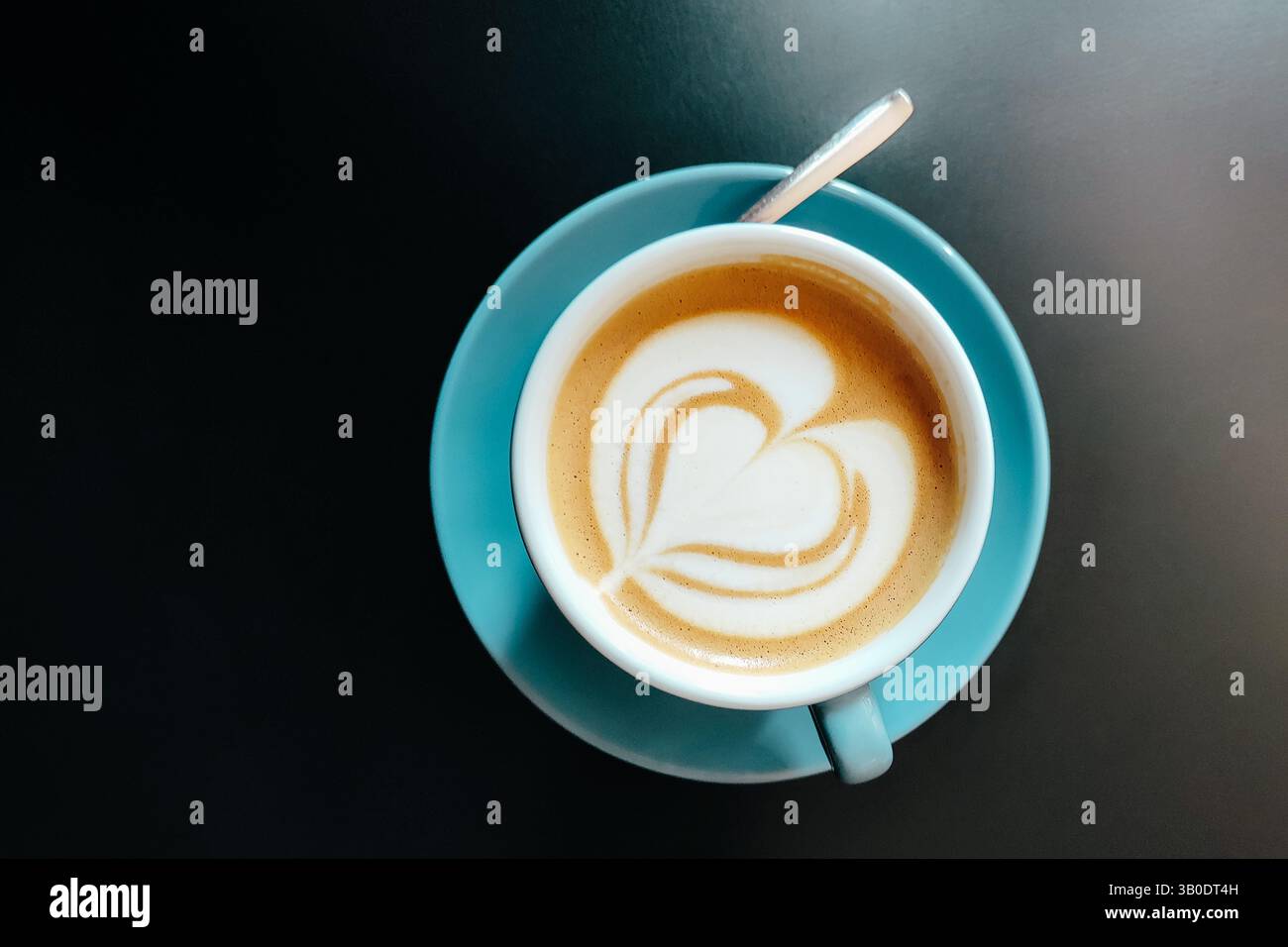 Tasse Cappuccino mit herzförmiger Latte Art auf Milchschaum in türkisfarbener Keramikschale mit Löffel auf dunklem Hintergrund. Draufsicht. Stockfoto