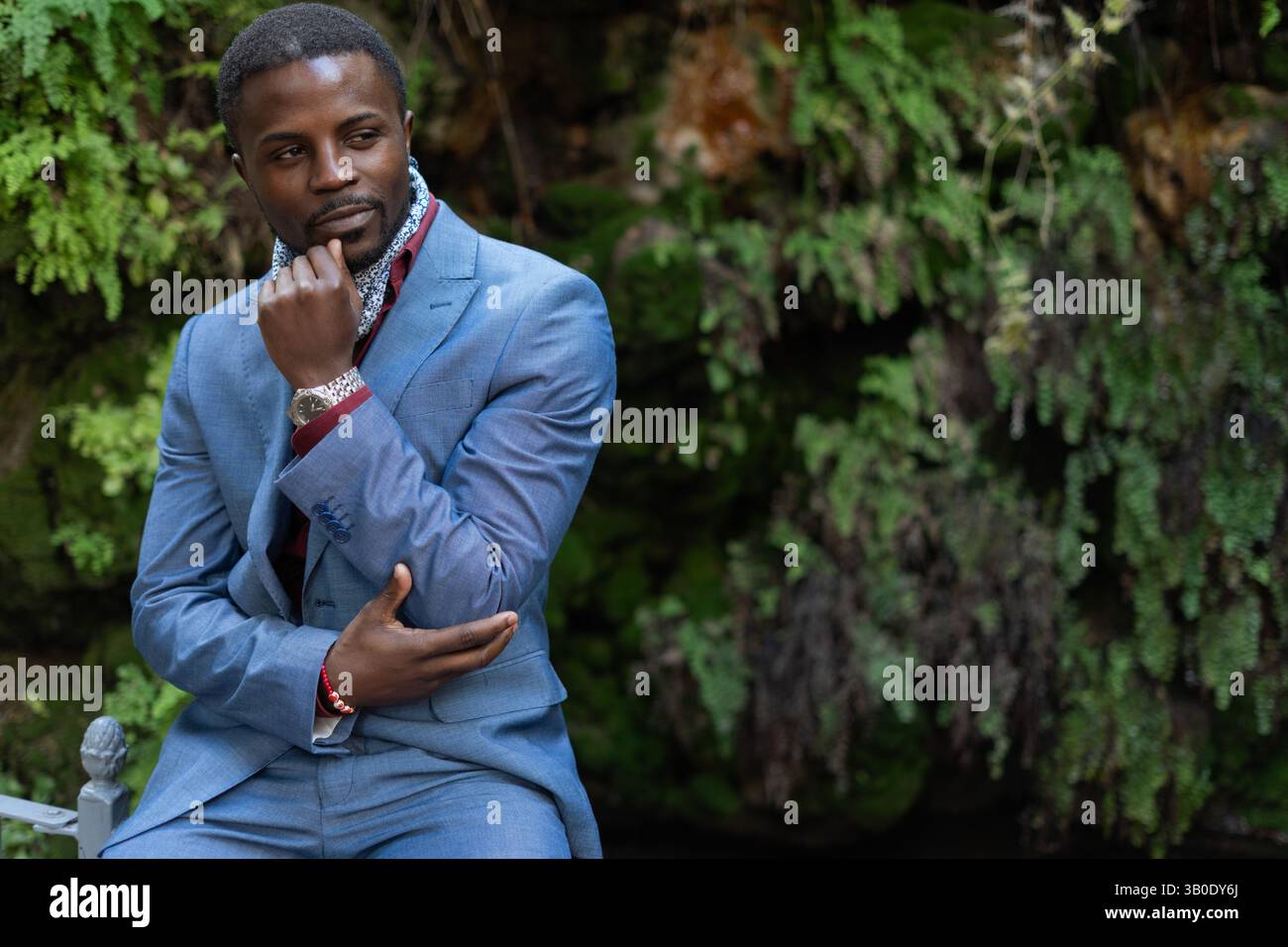 Nachdenklicher afrikanischer Geschäftsmann in blauem Anzug, sitzt in einem üppig grünen Park, berührt nachdenklich sein Kinn, während er verloren in die Ferne blickt Stockfoto
