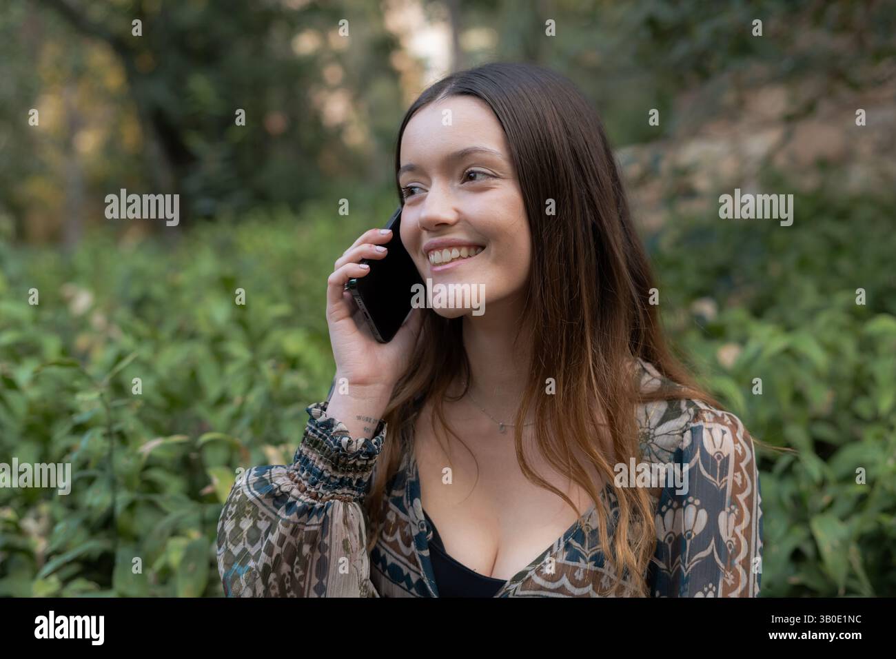 Glückliche junge Frau, die auf dem Smartphone spricht und in einem Park mit grüner Vegetation im Hintergrund lächelt Stockfoto