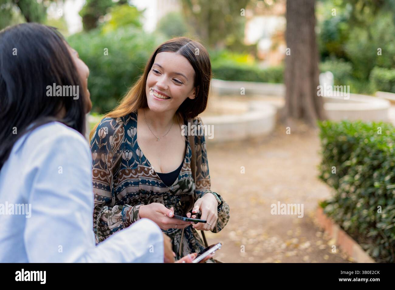 Zwei junge Frauen benutzen ihre Smartphones und lächeln, während sie auf einer Bank im Park sitzen und einen Moment der Entspannung und Verbindung genießen Stockfoto