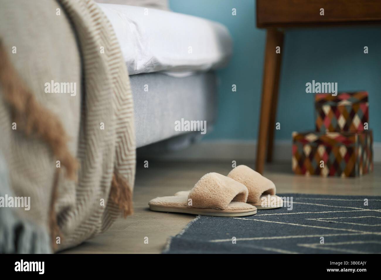 Komfortabel aussehendes Schlafzimmer mit weichen Hausschuhen, die auf Teppich neben Bett gelegt werden, und einem hölzernen Nachttisch mit farbenfrohen Kästen, die im Hintergrund sichtbar sind Stockfoto
