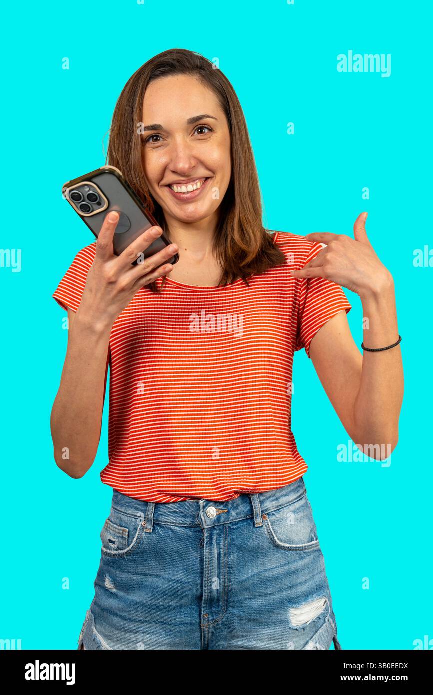 Eine junge Frau, die mit einem großen Lächeln auf türkisfarbenem Hintergrund auf ihr Smartphone zeigt Stockfoto