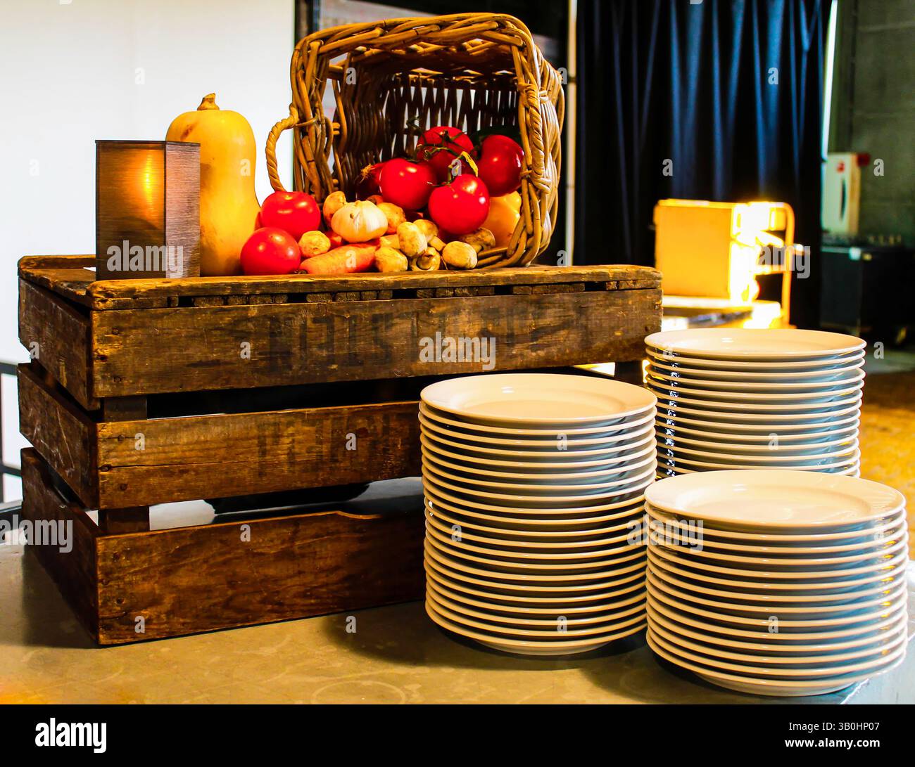 Eine rustikale Ausstellung mit einer Holzkiste gefüllt mit frischem Gemüse wie Tomaten, Kürbis und Pilzen. In der Nähe befindet sich ein Stapel weißer Platten Stockfoto