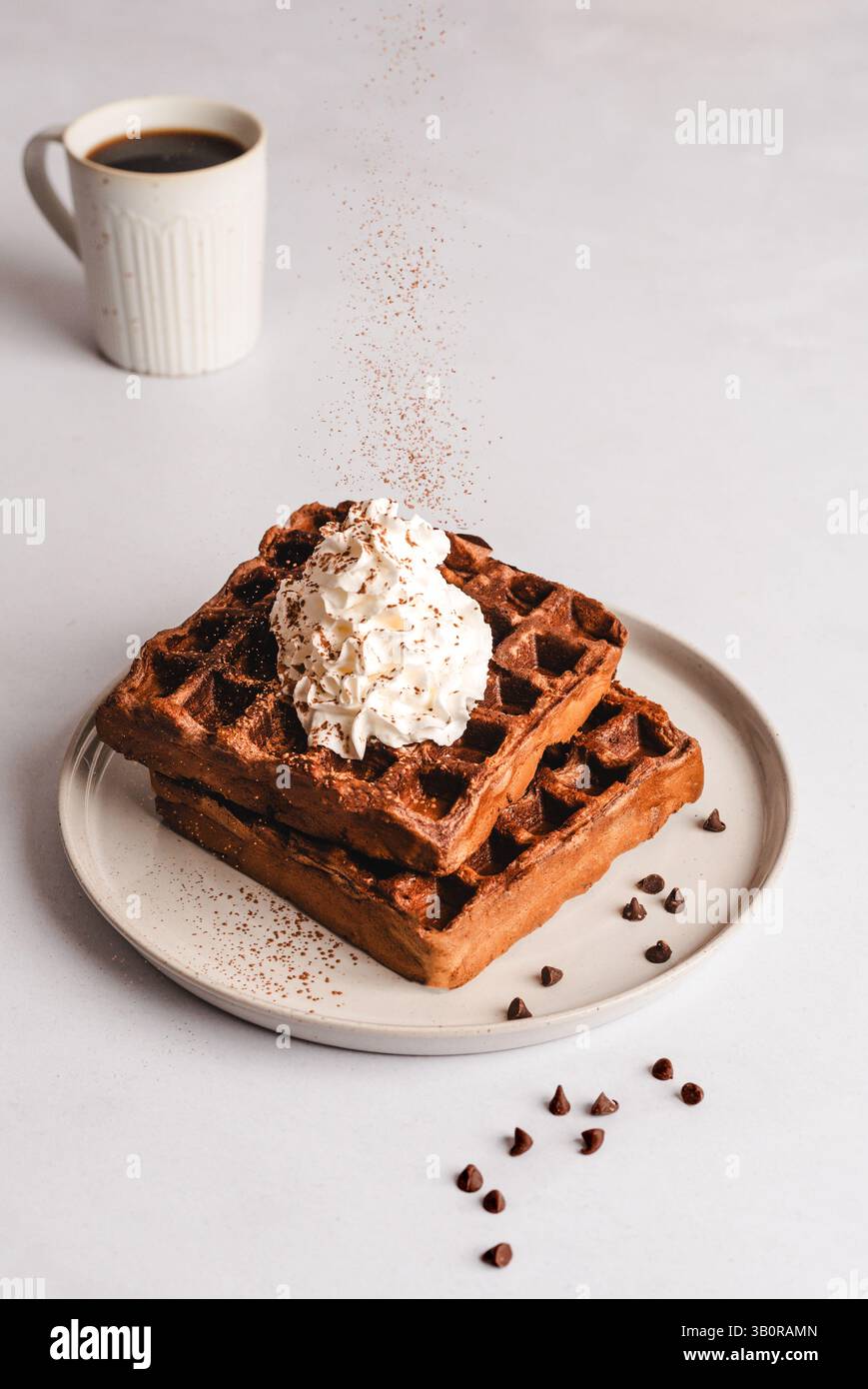 Ein dekadenter Stapel Schokoladenwaffeln, gekrönt mit Schlagsahne und mit Kakaopulver bestäubt, umgeben von verstreuten Schokoladenchips und Kaffee. Stockfoto