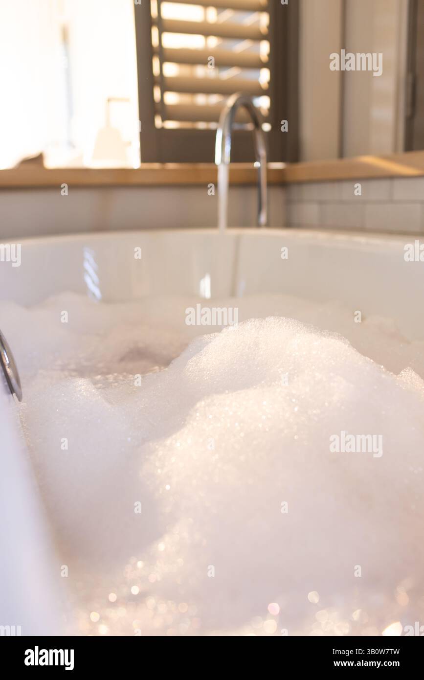 Die weiße Badewanne füllt sich mit schaumigen Seifenblasen in der Nähe des Chromhahns mit Holzjalousien Stockfoto