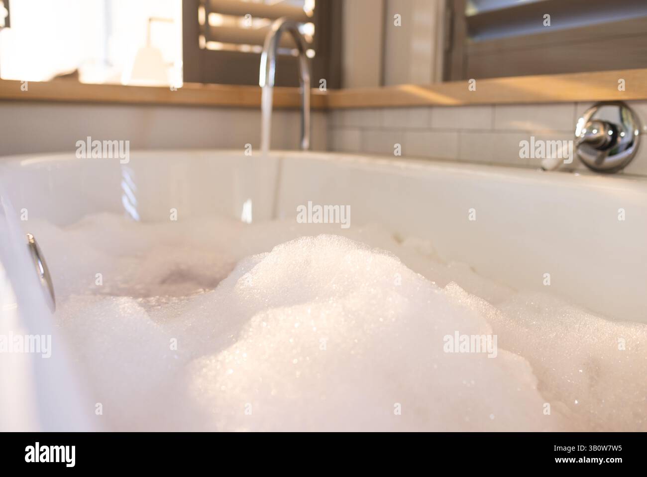 Die weiße Badewanne füllt sich mit Schaumblasen unter dem Chromhahn im Badezimmer, in der Nähe der Fensterläden Stockfoto