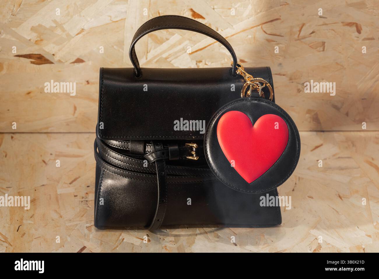 Mailand, Italien - 12. April 2025: Schwarze Moschino-Handtasche aus Leder mit rotem Schlüsselanhänger mit Herzanhänger. Designer-Accessoire auf hölzernem Hintergrund Stockfoto