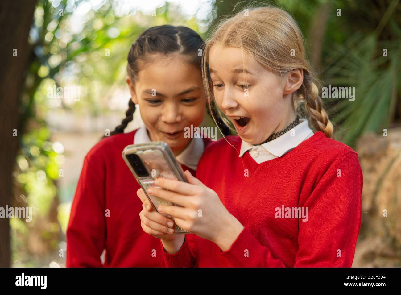 Zwei überraschte Schulmädchen in roten Uniformen, die in einem Park ein Smartphone anstarrten, in die digitale Welt eingetaucht und sich über das Staunen und die Freude ausstrahlten Stockfoto